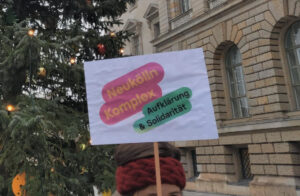 Schild mit der Aufschrift "Neukölln-Komplex. Aufklärung & Solidarität" vor Weihnachtsbaum und Gebäude des Abgeordnetenhauses